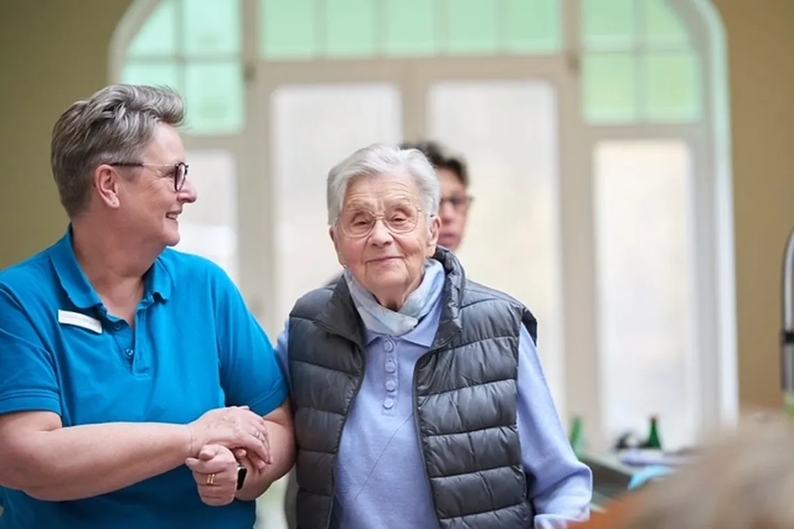 Eine Mitarbeiterin und eine Seniorin stehen Hand in Hand beieinander und lächeln.