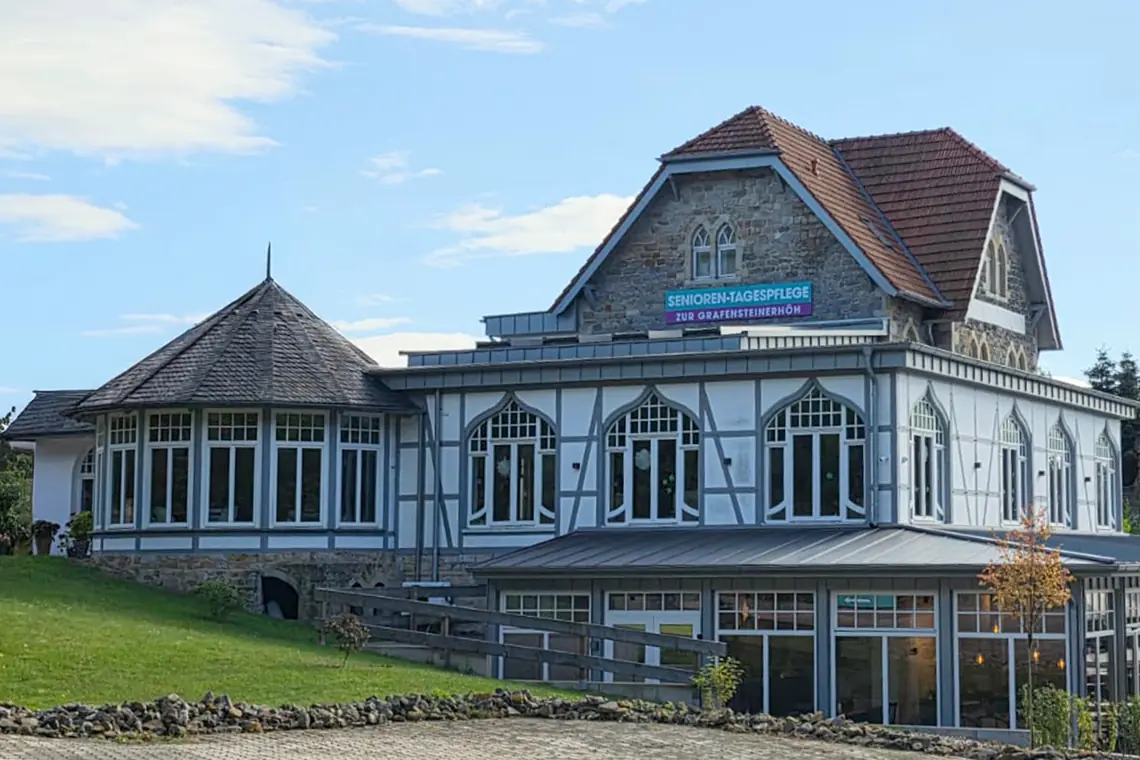 Das historische Haus „Zur Grafensteinerhöh“ in Rinteln: Fachwerkfassade mit spitzbogigen Fenstern, Eckturm und rotem Ziegeldach, darüber das Schild der Seniorentagespflege; davor Rasen und der gepflasterte Vorplatz.