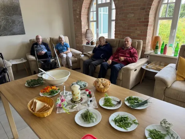 Ein gedeckter Tisch mit frisch zubereiteten Salaten, dahinter Gäste in der Sitzgruppe.