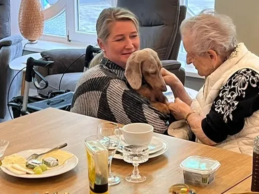 Eine Mitarbeiterin und eine Seniorin am gedeckten Tisch, auf dem Schoß einer der beiden Dackel.